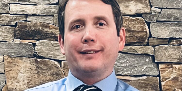 Chris calling standing in front of a rock wall with a blue button down and tie