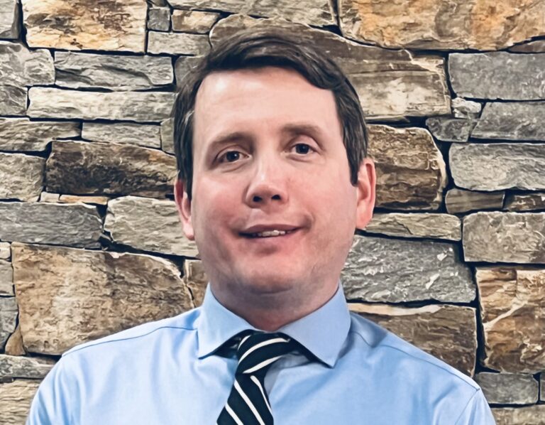 Chris calling standing in front of a rock wall with a blue button down and tie