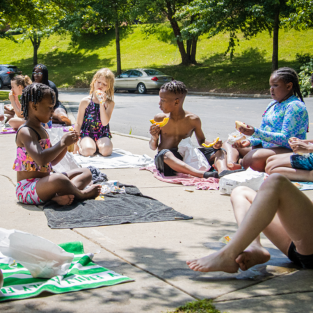 young people sitting on beach towels on sidewalk eating fruit