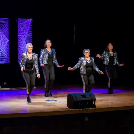 four ladies wearing black and silver dancing on stage with purple banners in the background
