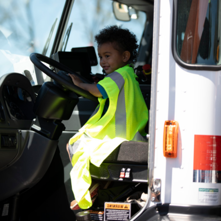 young boy sitting in drivers seat of big truck, wearing safety vest