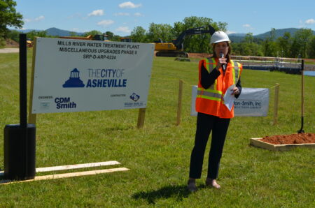 Mayor Manheimer holding microphone to address crowd beside a Mills River plant / city of Asheville sign