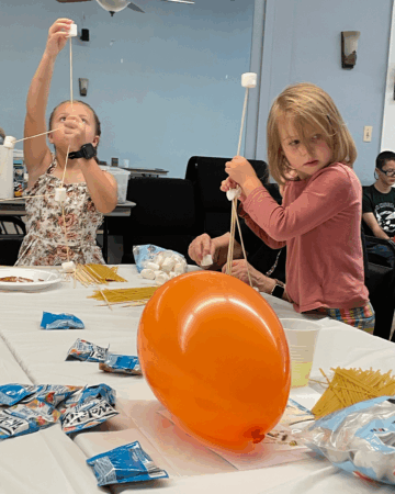 two young girls building with spaghetti noodles, marshmallows and balloons