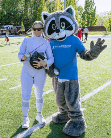 woman wearing fencing outfit stands with parks and recreation raccoon mascot
