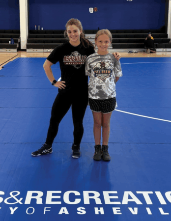 a teenage girl and a younger girl stand on wrestling mat in a gym