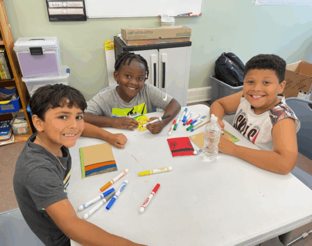 three young boys sit at a table with magic  markers and paper booklets