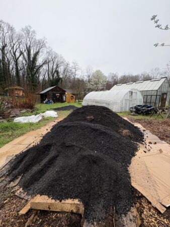 a pile of black biochar material in a grassy area near greenhouse buildings.
