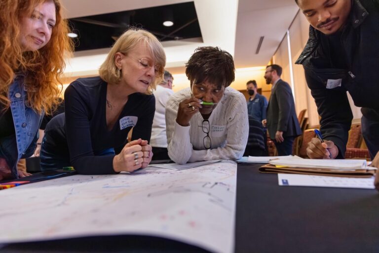 Four adults lean over a large printed map on a table, studying it closely and marking notes during a community planning session.